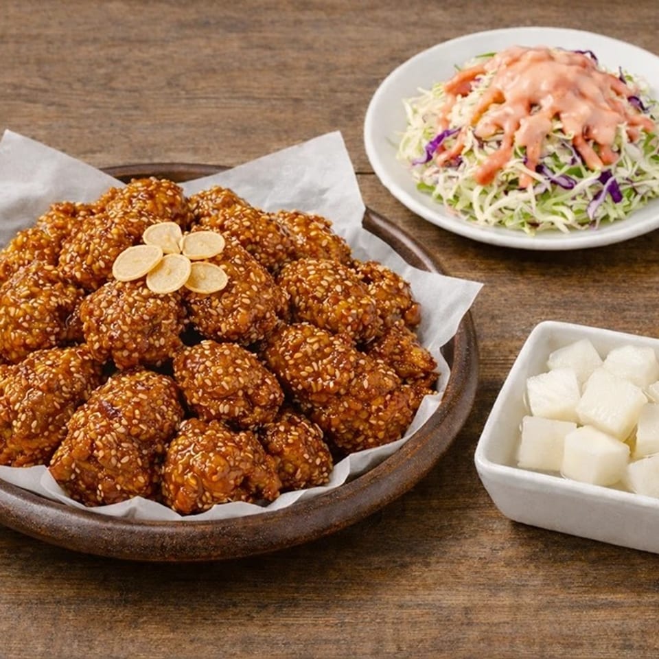 Plate of sesame-coated fried chicken with garlic slices, cabbage salad, and cubed pickled radish on a wooden table.