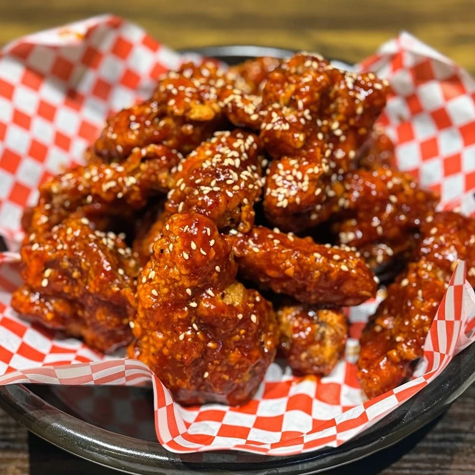 Glazed chicken wings sprinkled with sesame seeds, served in a black basket lined with red-and-white checkered paper.